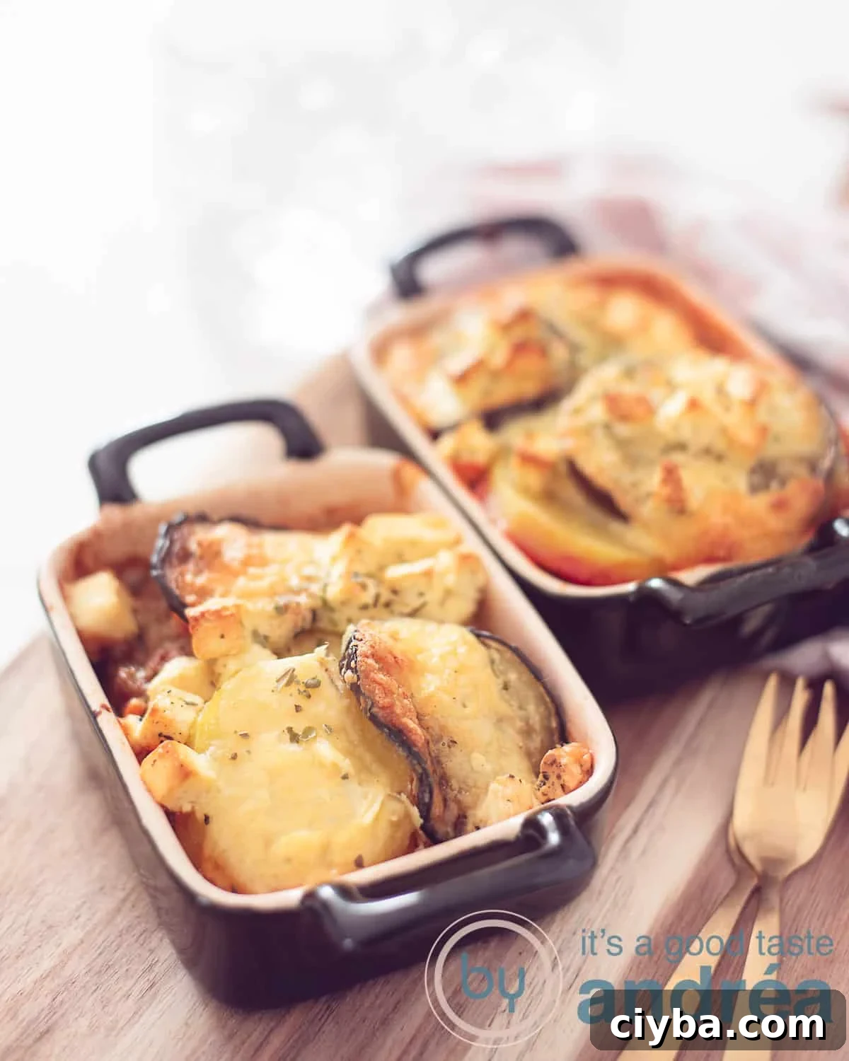 An elevation photo with two black oven dishes on a black shelf filled with Moussaka from Greece. Right front two forks.