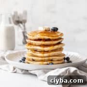 action shot of syrup being poured onto a stack of blueberry muffin mix pancakes topped with a pad of butter and extra fresh blueberries.