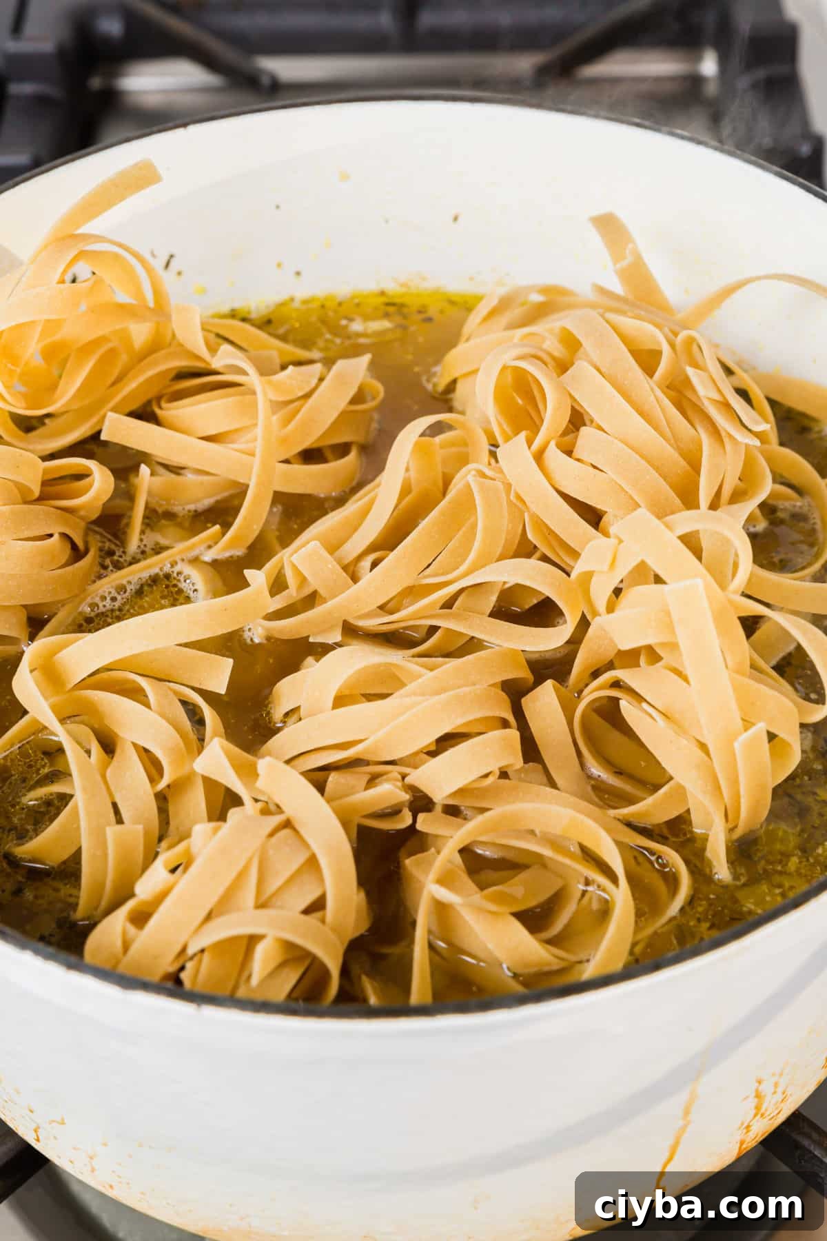 A white dutch oven of gluten free egg noodles added to a pot of chicken noodle soup before the noodles have been cooked.