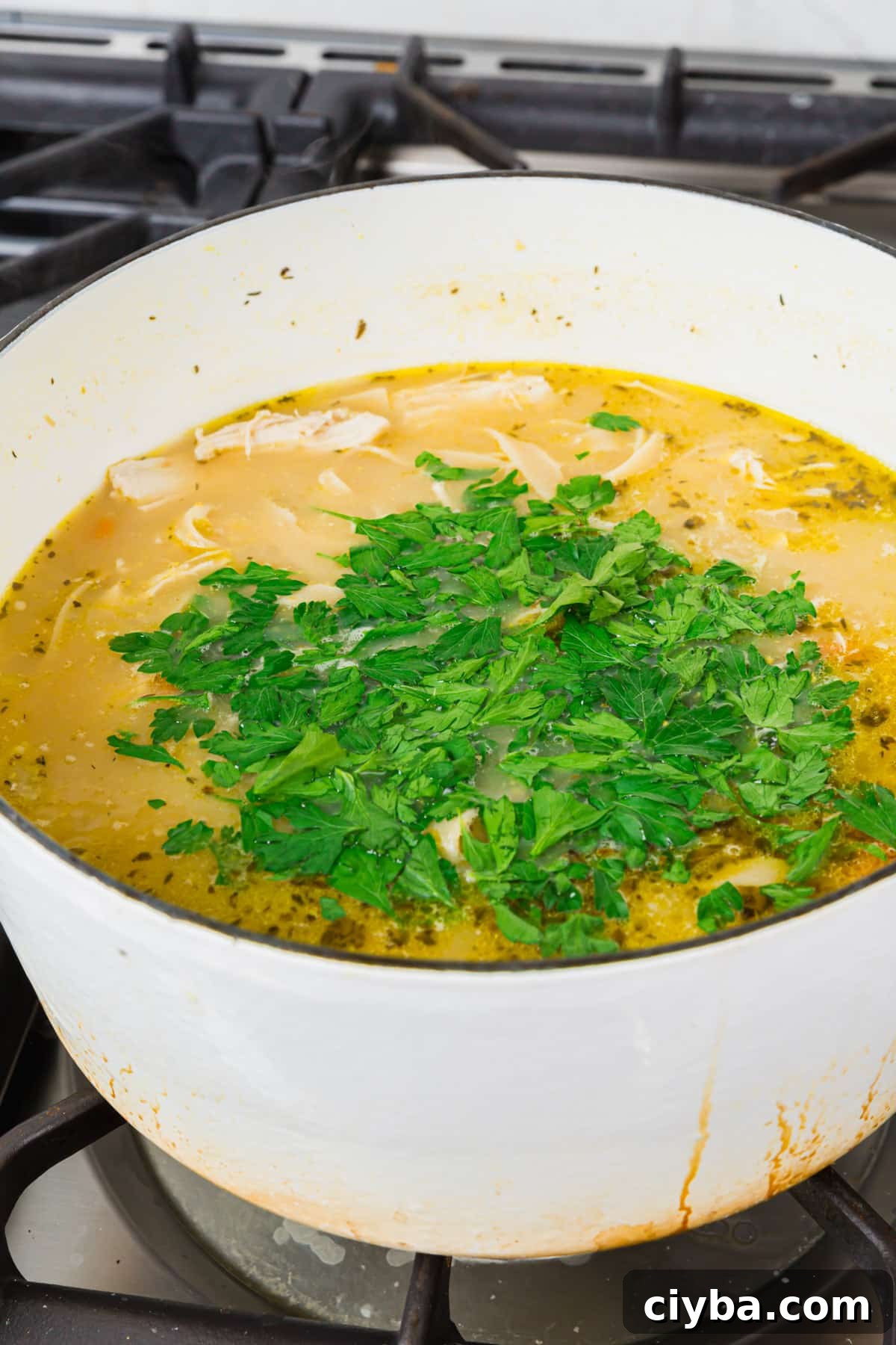 A white dutch oven on the stove with gluten free chicken noodle soup in it topped with a pile of chopped fresh parsley before mixing together.