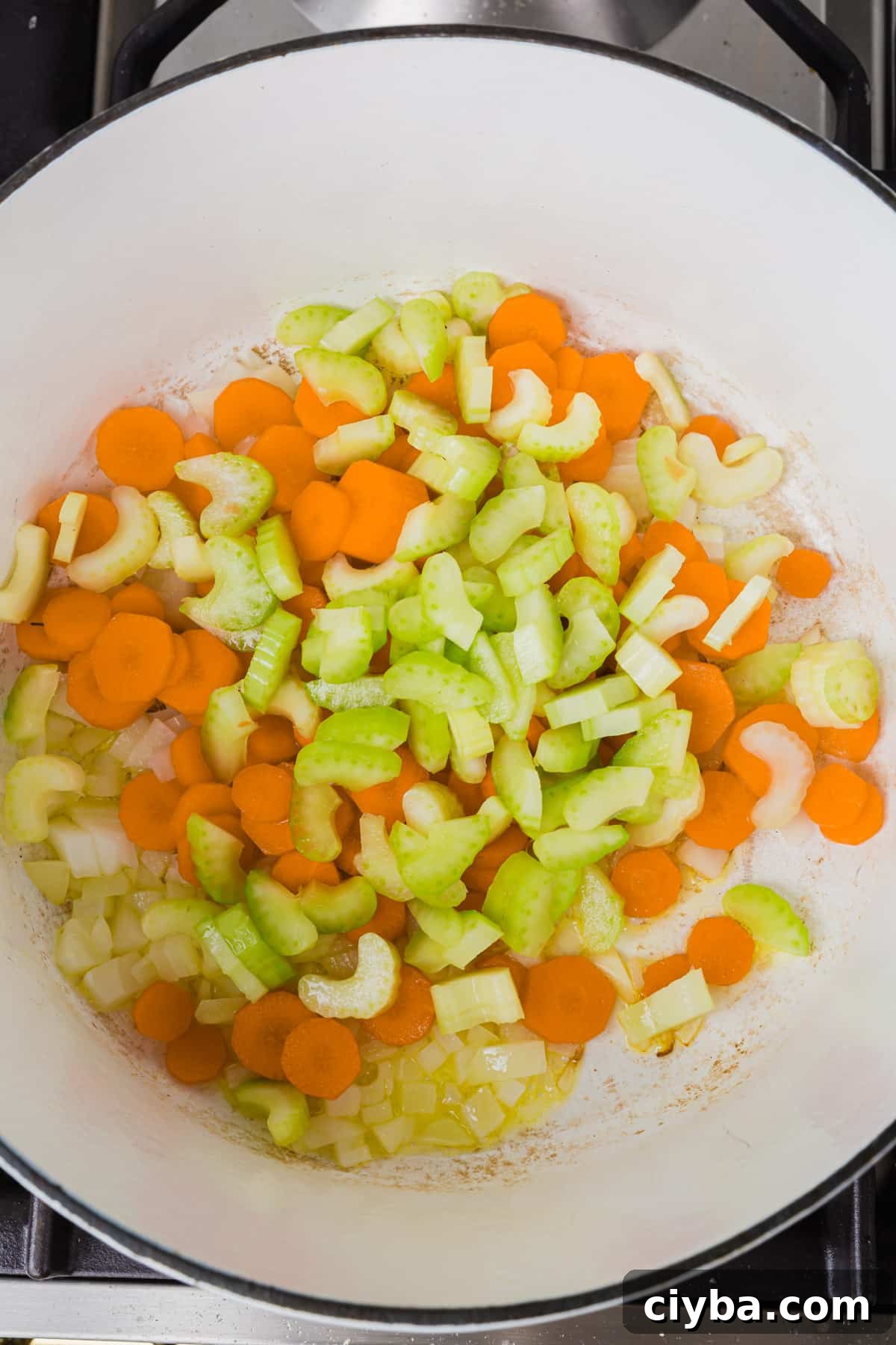 An overhead view of a white dutch oven with raw diced celery, carrots and onion in olive oil before cooking.