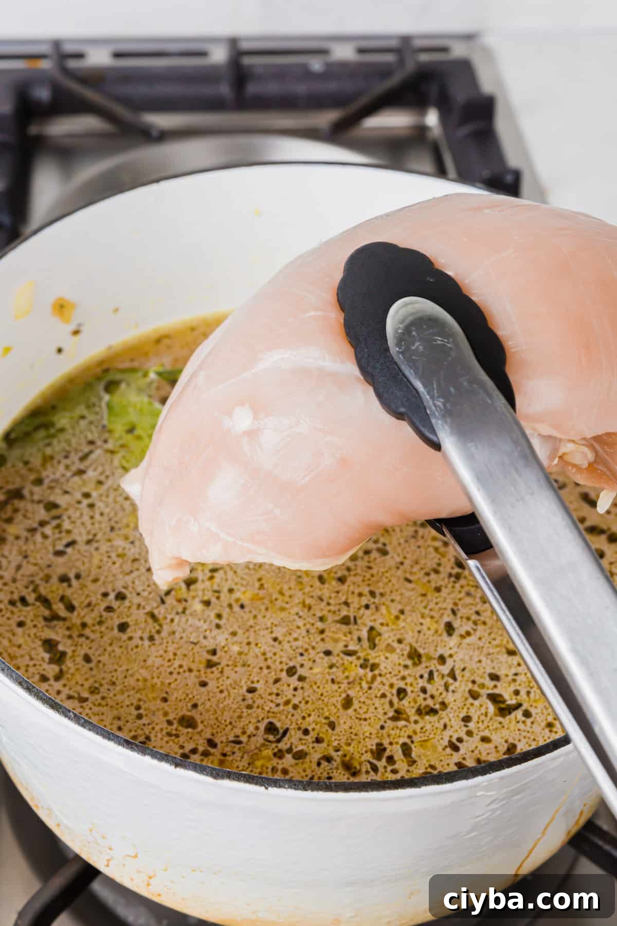 A pair of tongs lowering a raw chicken breast into a dutch oven of chicken broth, herbs, bay leaves, and minced garlic.