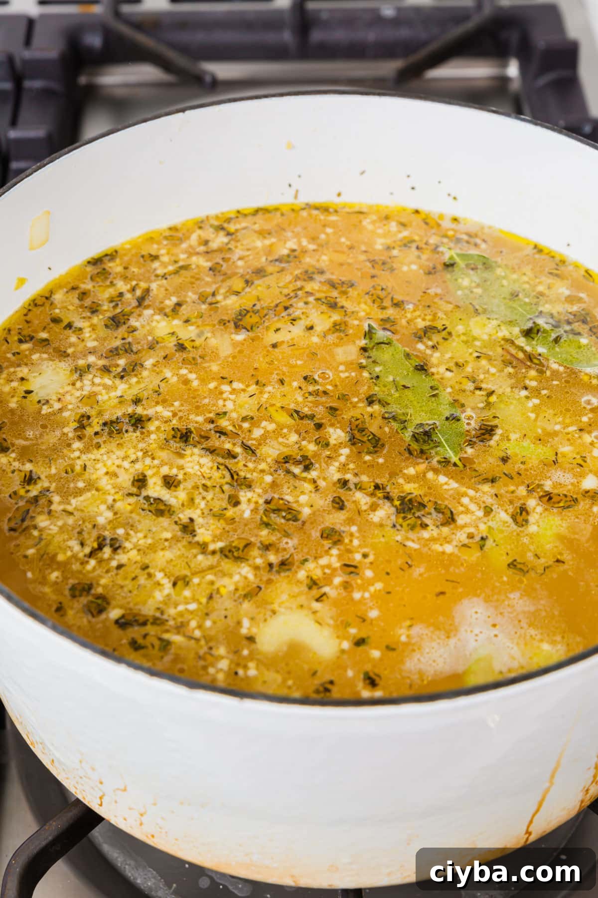 A white dutch oven of chicken broth, spices, garlic, and bay leaves simmering on the stove top.