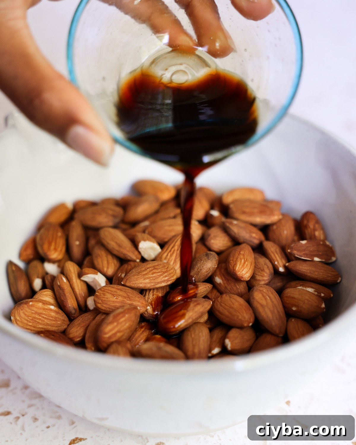Drizzling a small dish of tamari over hot roasted almonds in a white bowl.
