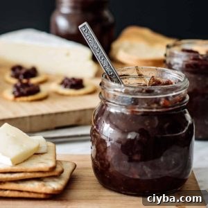 a jar with red onion chutney with a spoon inside, with crackers and cheese on the side, over a wooden board.