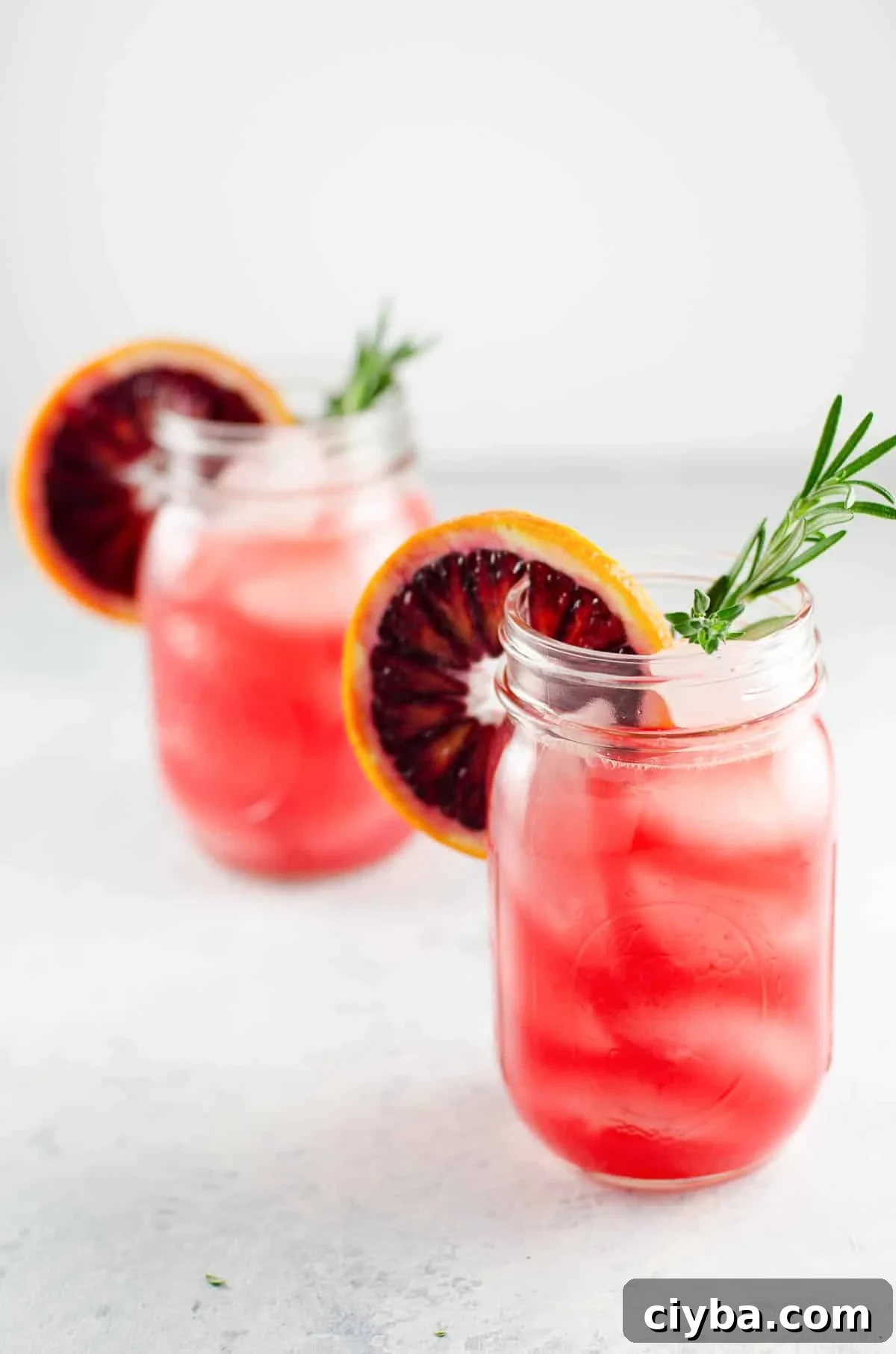 two blood orange cocktails in mason jars, each garnished with a rosemary sprig and a blood orange wheel
