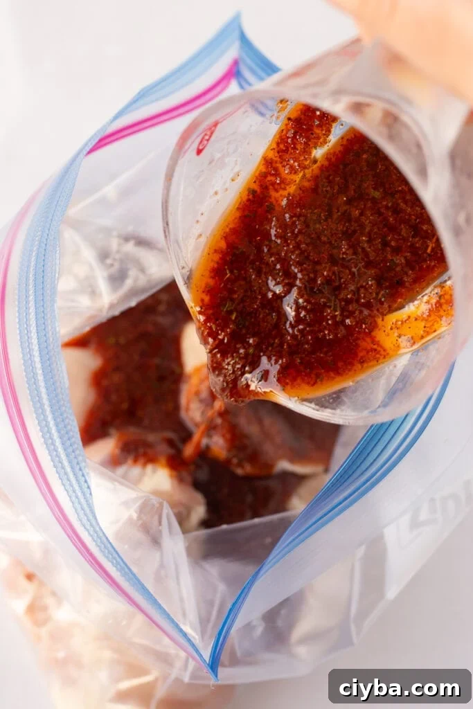 Close up of a red marinade being poured into a gallon sized ziploc bag with raw boneless skinless chicken thighs in it.
