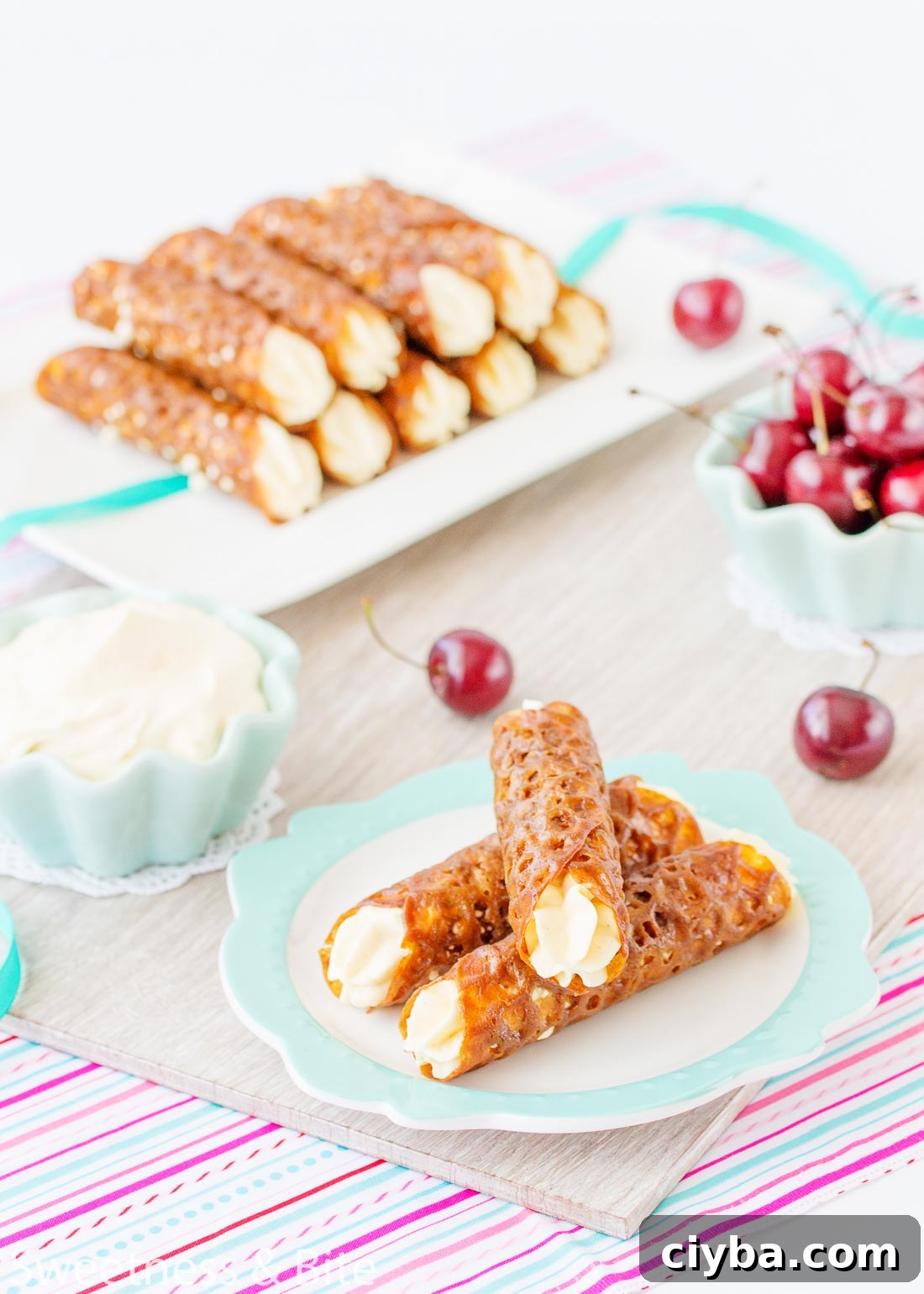 Three gluten free brandy snaps stacked on a plate in the foreground, with a bowl of whipped cream and fresh cherries in the background.