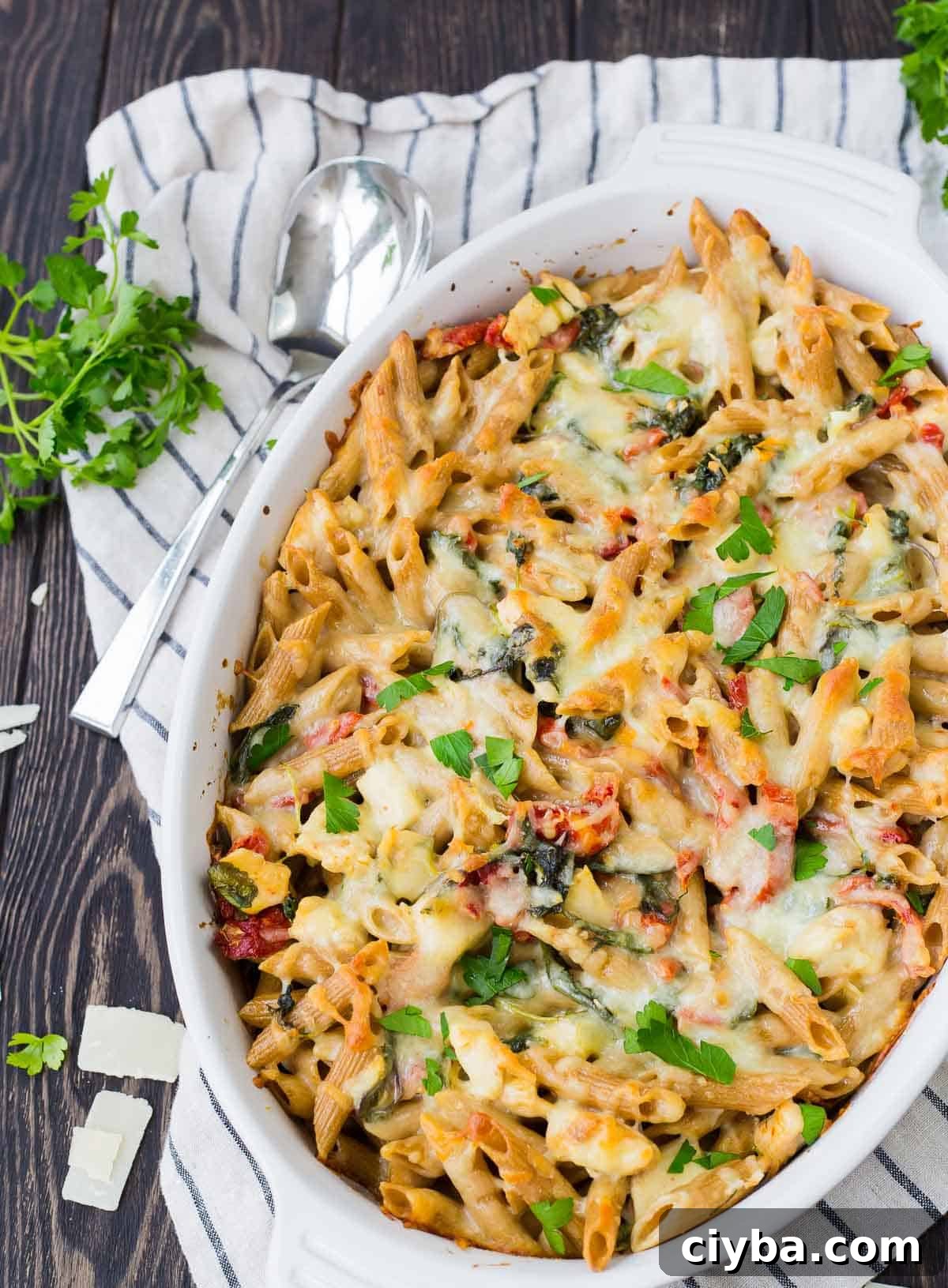Overhead of baked pasta in white casserole dish, with spoon alongside, on striped cloth.