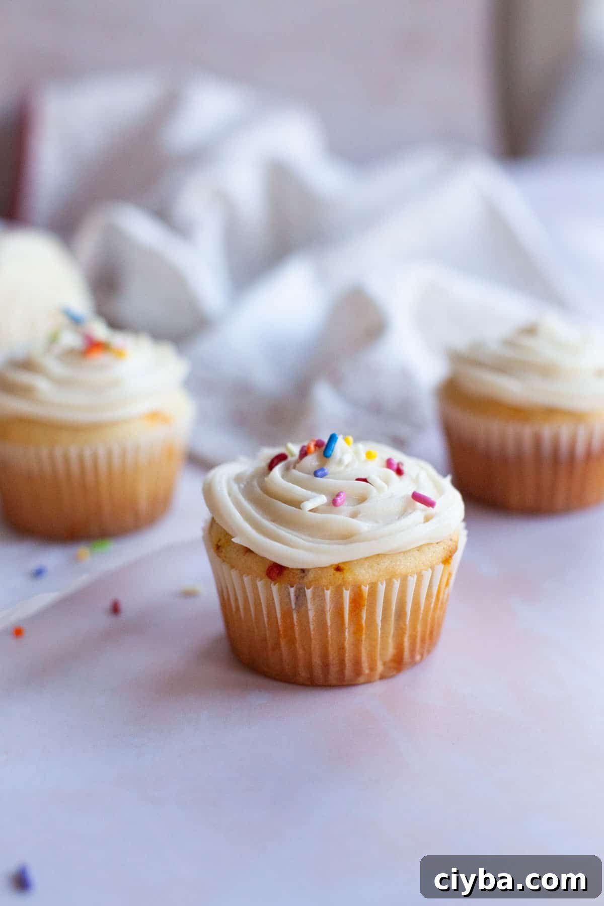 Confetti Cupcakes with sprinkles on top on a pink countertop with more cupcakes and sprinkles.
