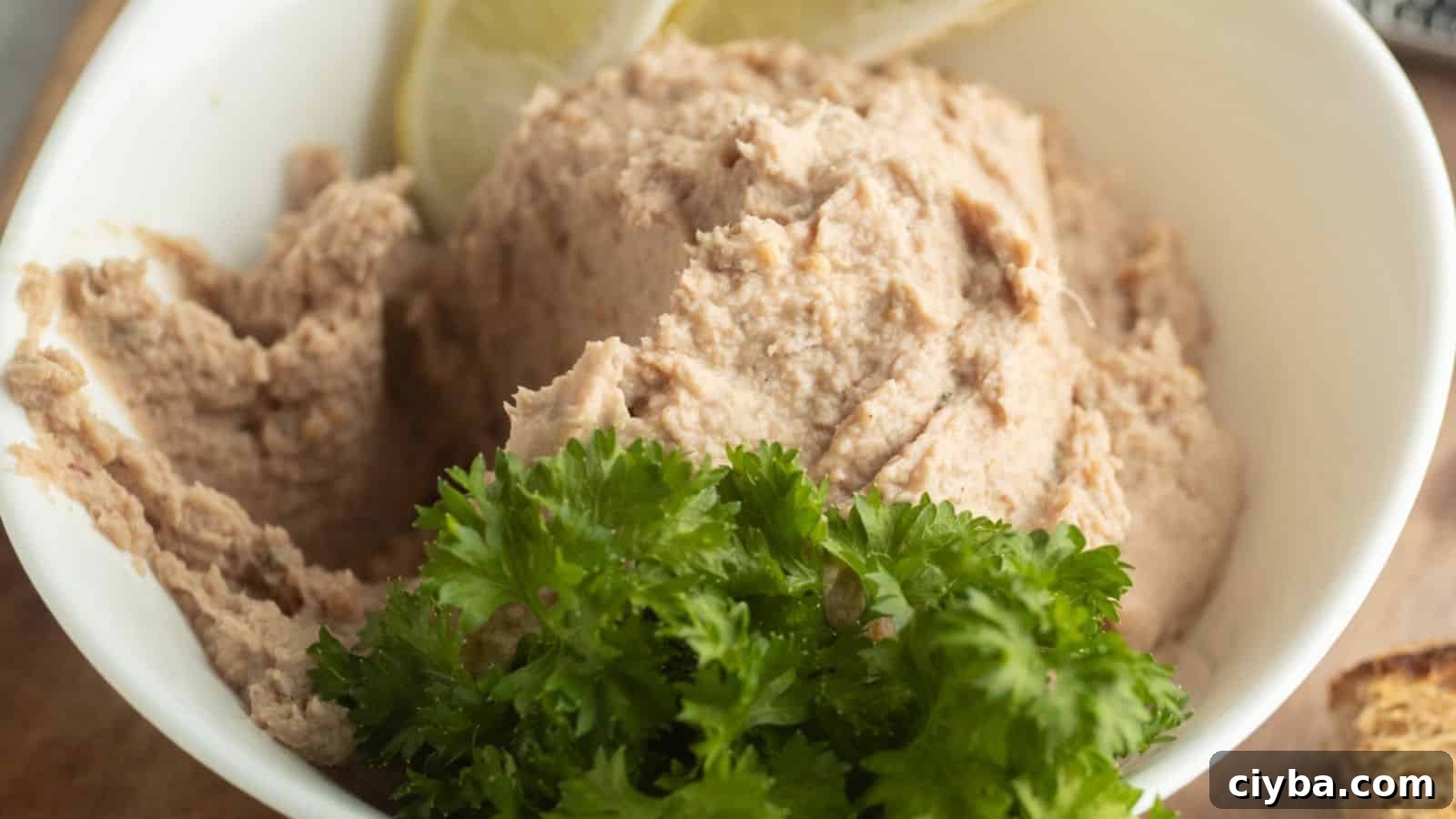 A white bowl filled with a creamy, light brown spread, garnished with fresh parsley and lemon slices.