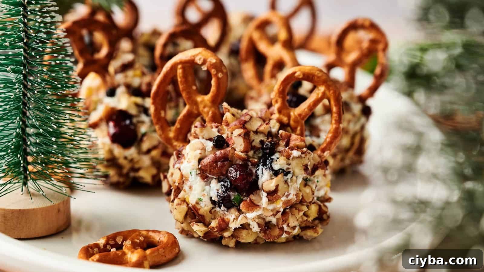 Cheese balls coated with chopped nuts, topped with cranberries and pretzels, arranged on a white plate near a small artificial pine tree.