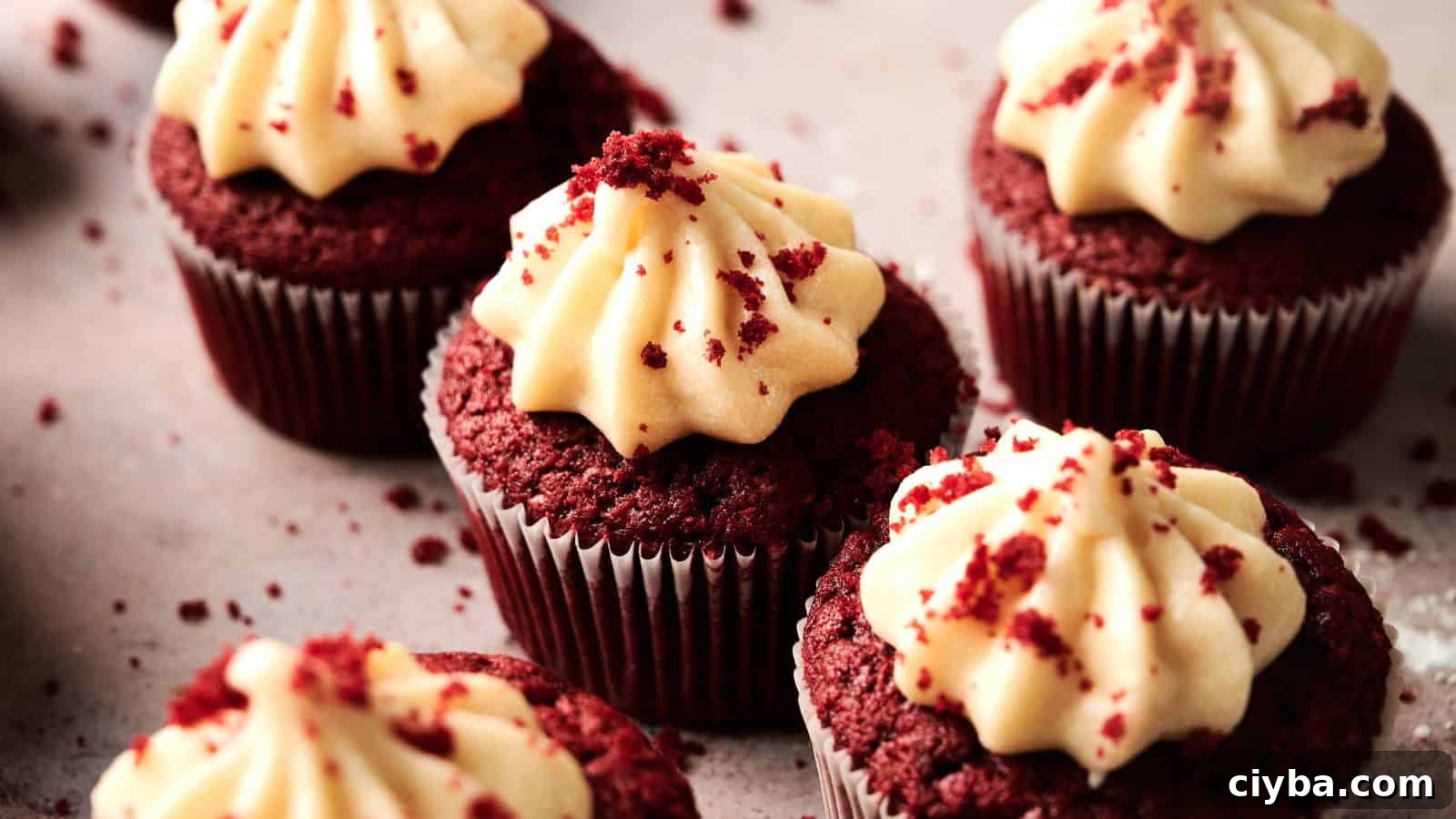 Several red velvet cupcakes with swirls of cream cheese frosting and sprinkled crumbs on top, arranged on a light-colored surface.
