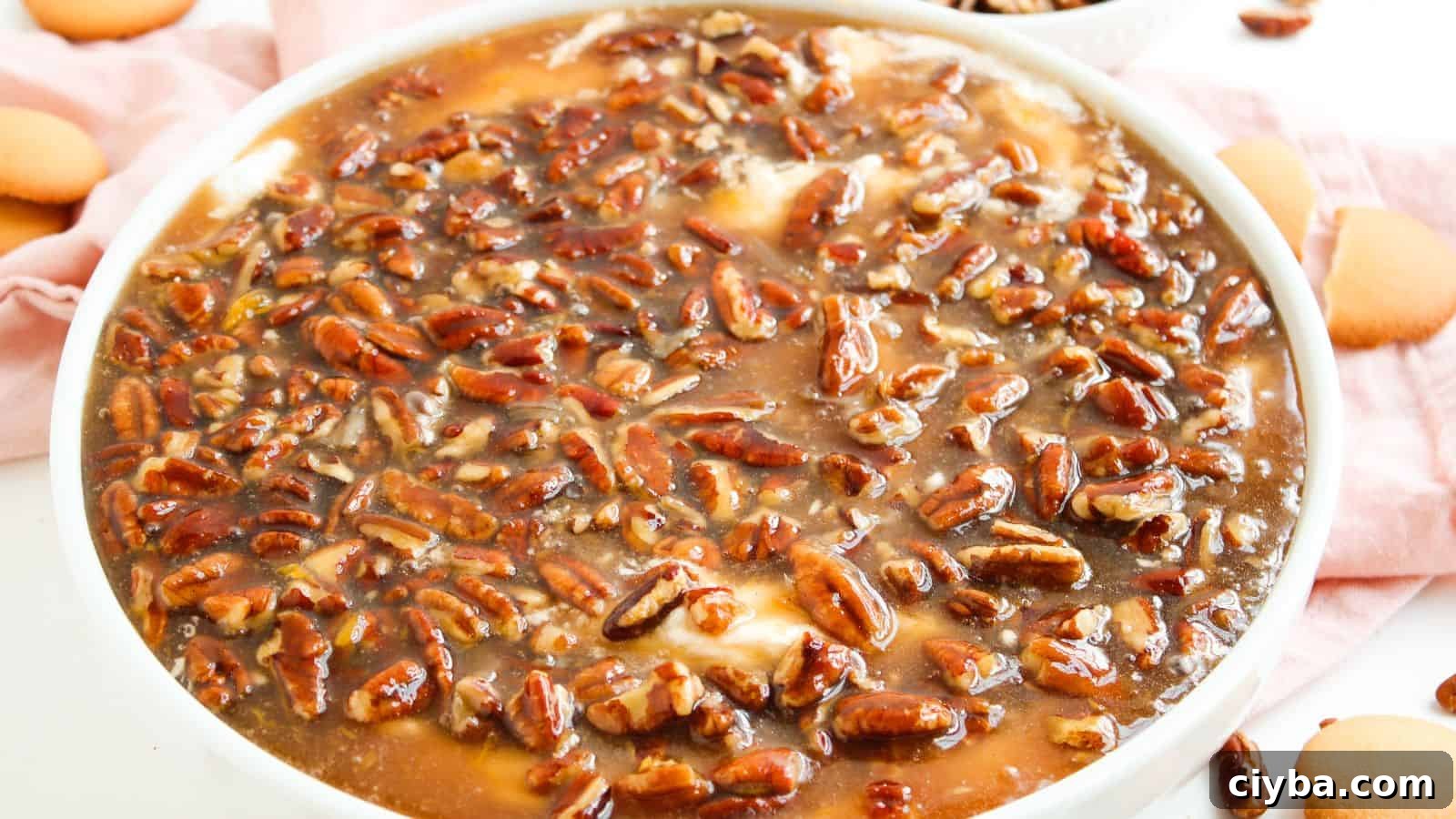 A close-up of a dessert topped with a glossy layer of pecans and caramel sauce in a white dish, with vanilla wafers on the side.