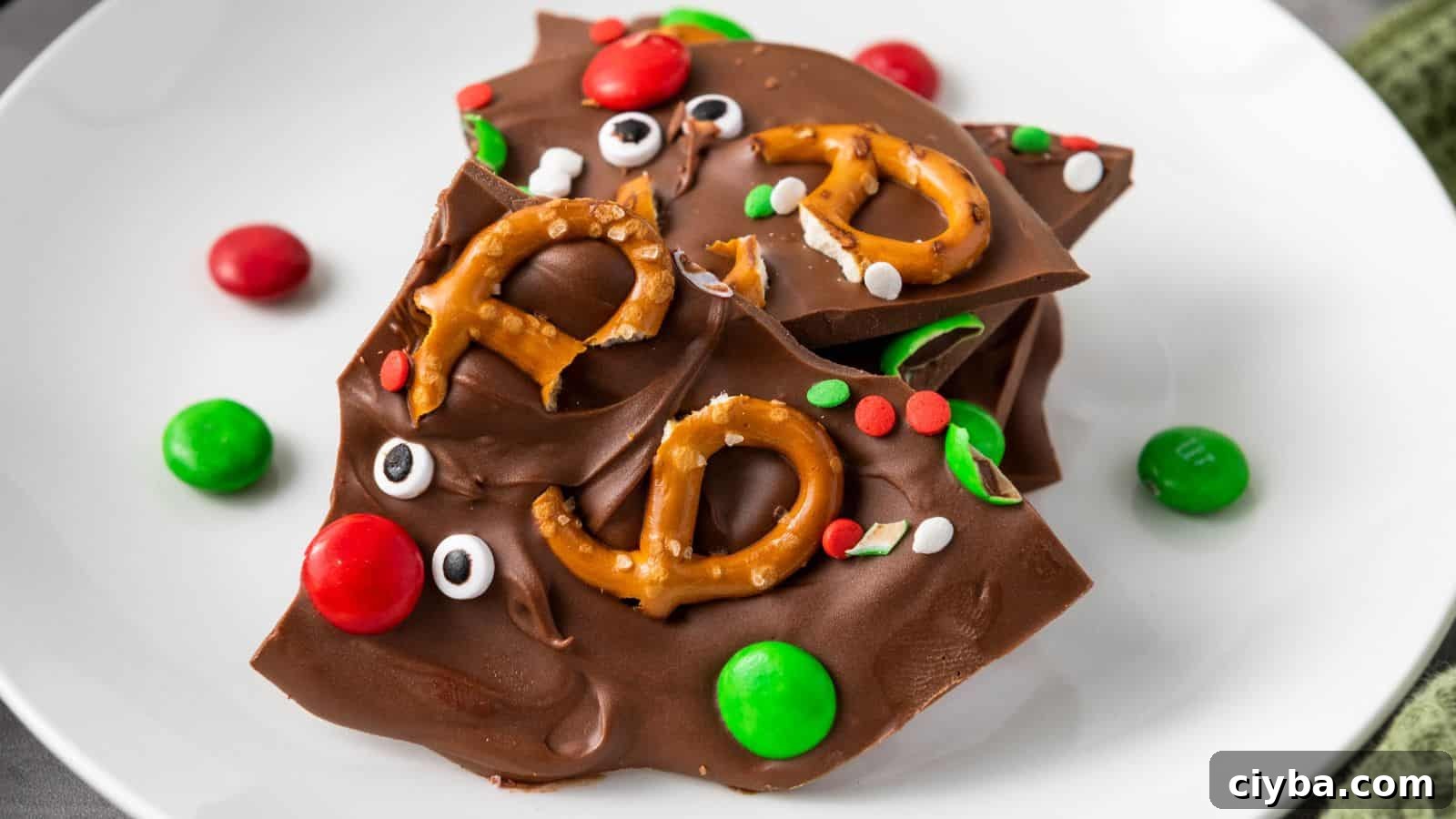 Chocolate bark topped with mini pretzels, red and green candy-coated chocolates, candy eyes, and round sprinkles on a white plate.