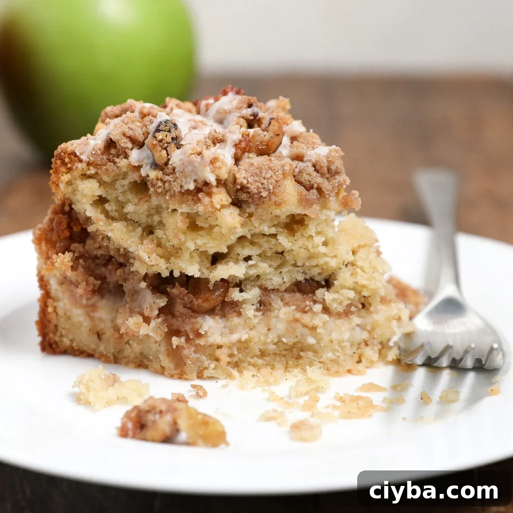 Cream Cheese Apple Coffee Cake Recipe for Fall Baking 7 A slice of Apple Coffee Cake with Cream Cheese sits on a white plate with a fork beside it. The cake has a crumbly streusel layer and is topped with icing. A green apple is blurred in the background.