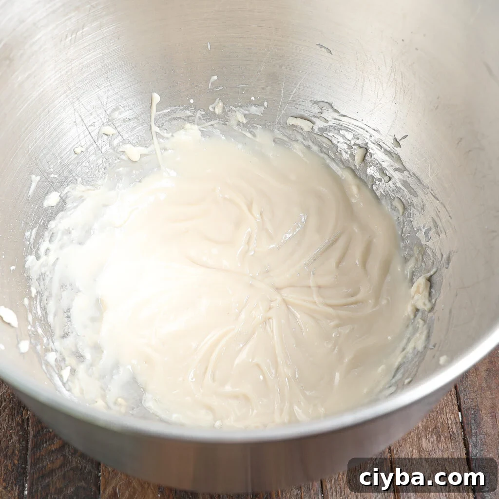 Cream Cheese Apple Coffee Cake Recipe for Fall Baking 3 A metal mixing bowl containing a smooth, creamy white batter or frosting—perfect for an Apple Coffee Cake with Cream Cheese—partially spread around the sides, sits on a wooden surface.