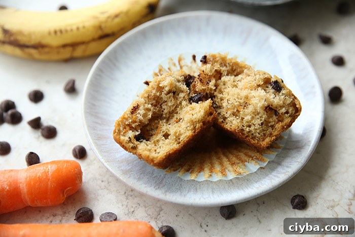 A torn in half banana carrot chocolate chip muffin sitting on the muffin wrapper on a dessert plate. There's a very ripe banana in the upper left corner, two carrots in the bottom left corner, and chocolate chips scattered around the small plate.