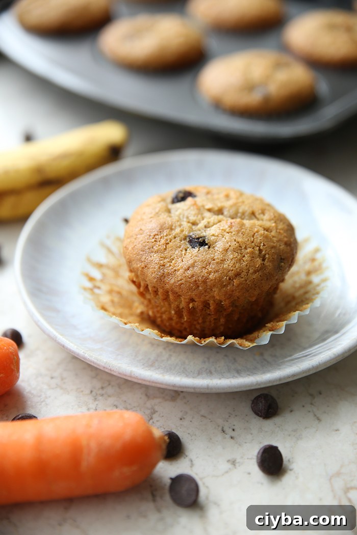 An unwrapped carrot banana chocolate chip muffin sitting on the wrapper on a small plate. Faded in the upper background is part of a muffin pan of muffins and an overripe banana. The tip of a carrot and half a carrot are in the bottom left corner.