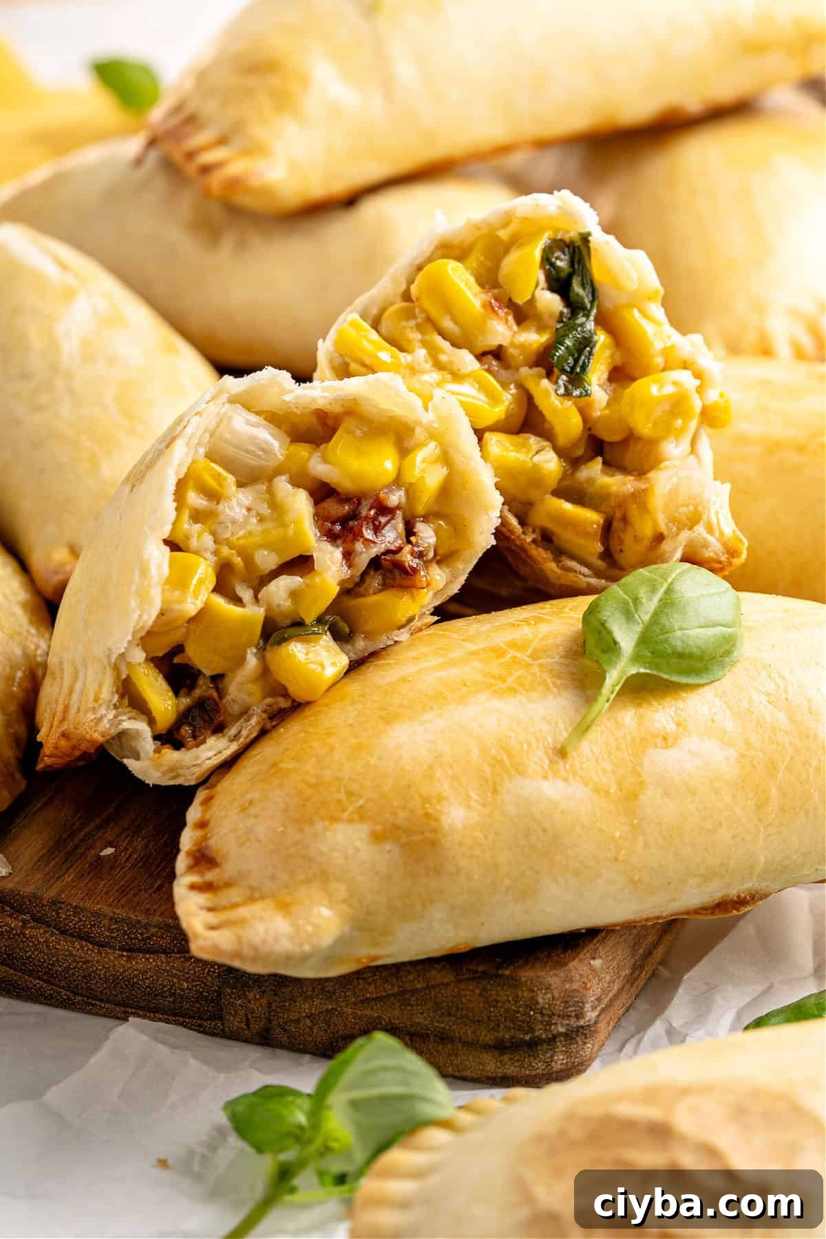Two corn empanada halves on whole ones. Basil leaves. Wooden board.