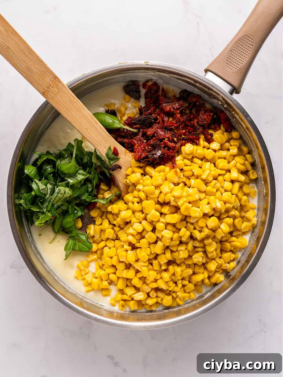 Basil, dried tomatoes and corn added to white sauce in metal pot.