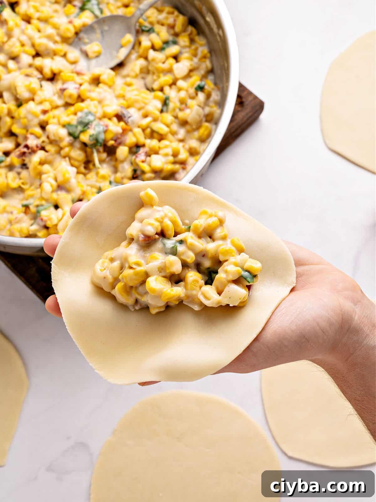 Holding empanada disc with corn filling over skillet and white background.