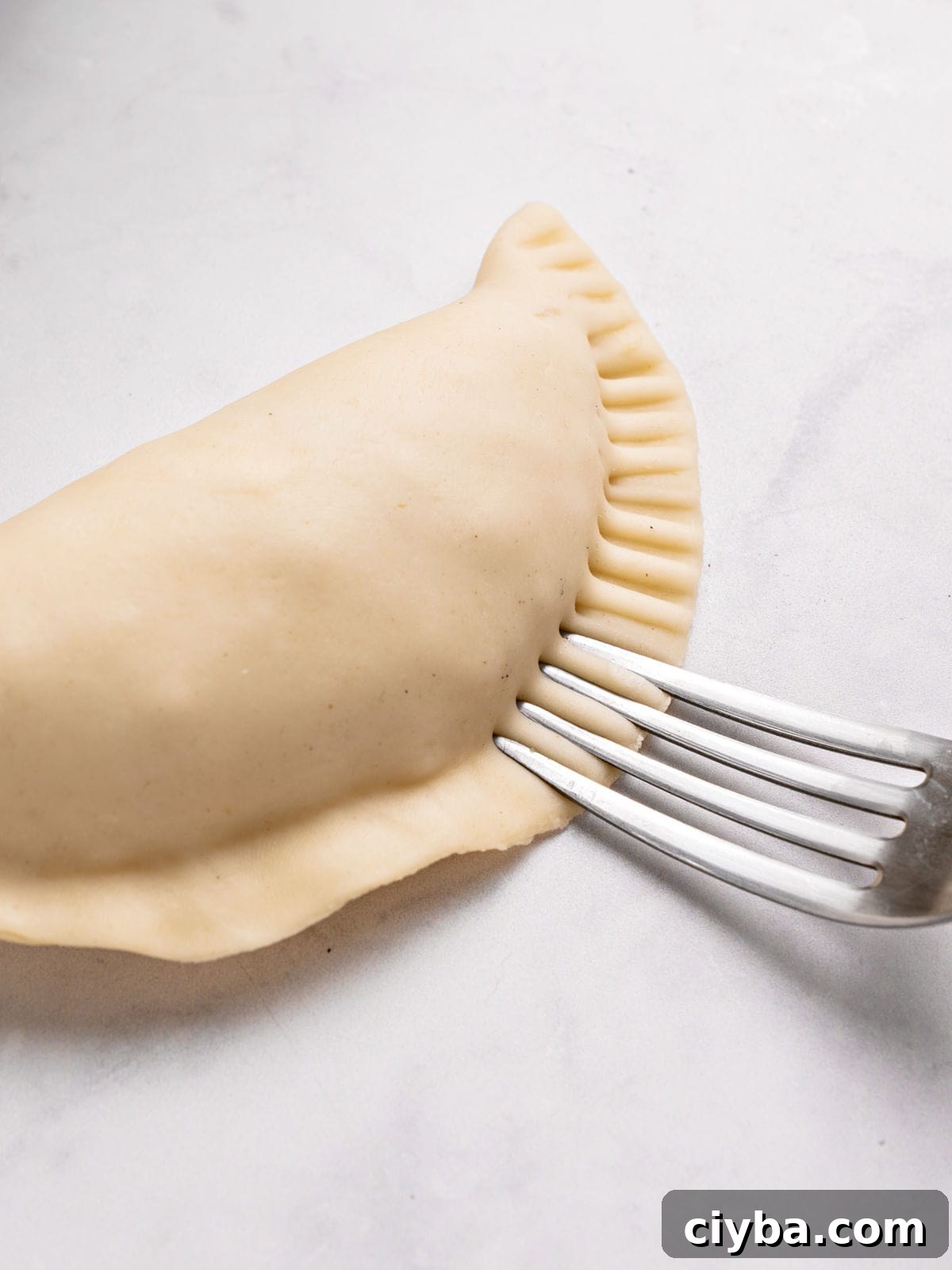 Crimping an empanada with a silver fork. White sauce with onion in a metal saucepan. White background.