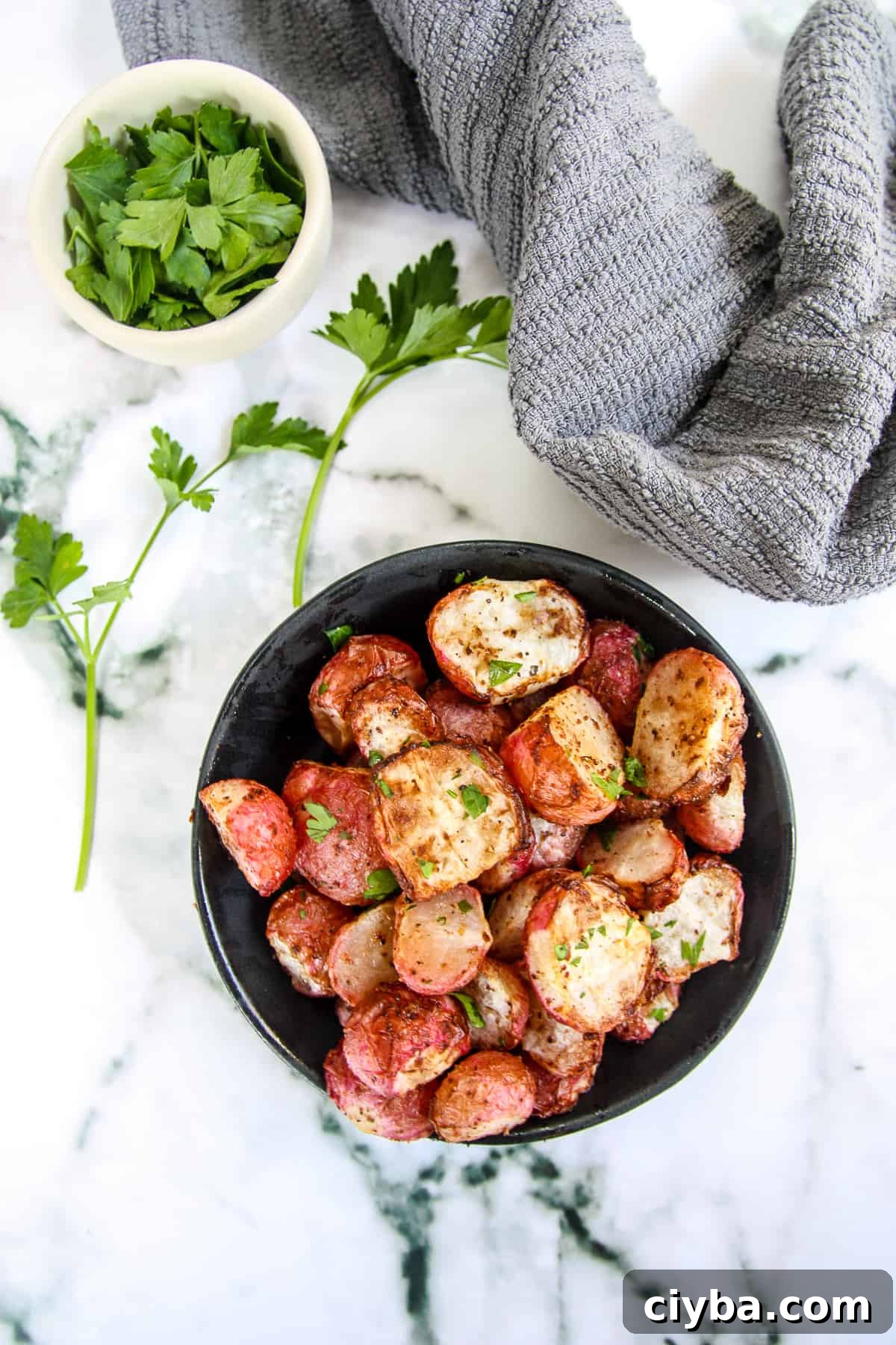 A black bowl filled with air fryer radishes next to a small white dish of parsley and a gray kitchen towel.