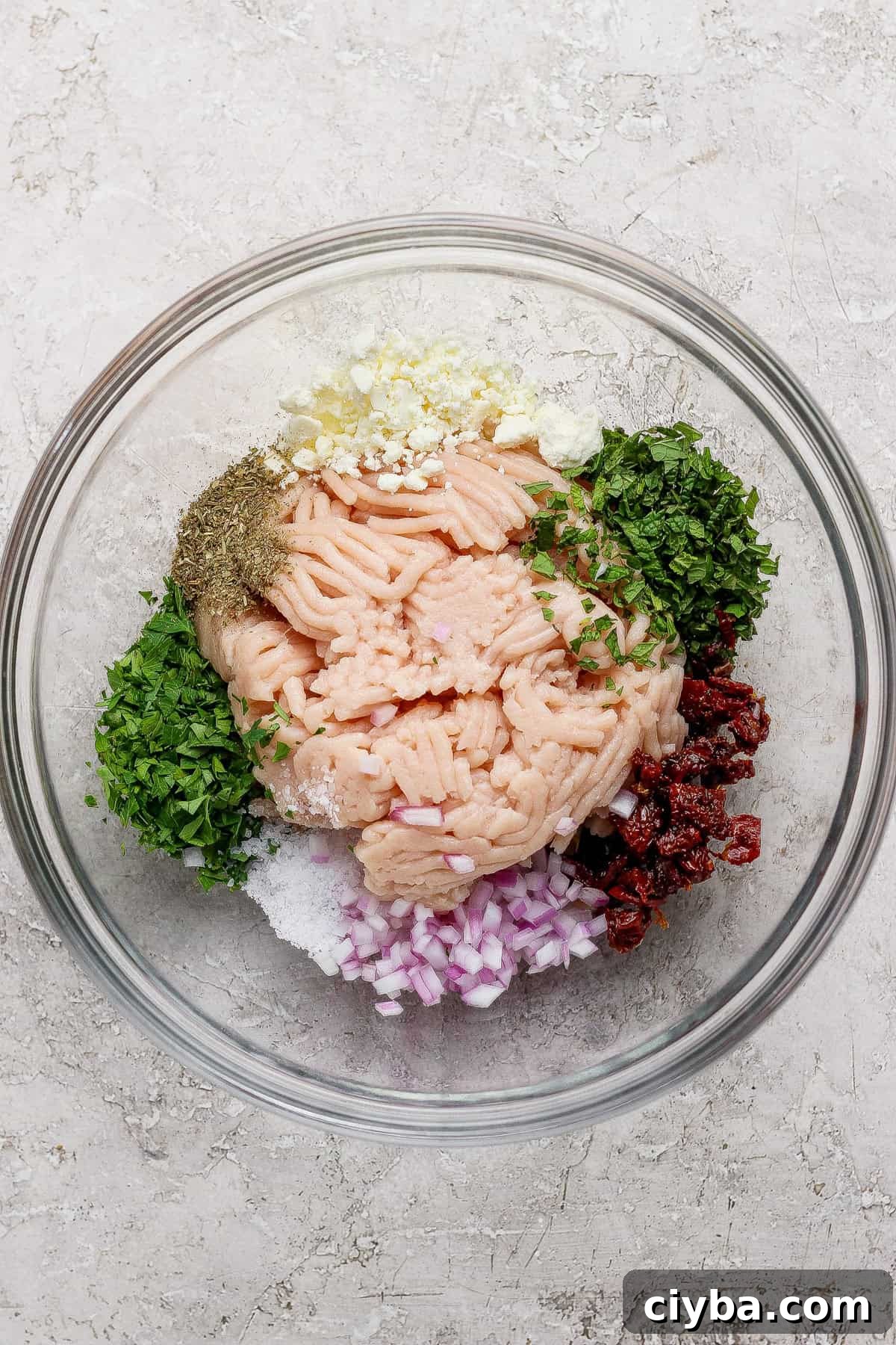 Crispy Air Fryer Mediterranean Chicken Bites with Herbs 3 A glass bowl containing ground meat, chopped red onion, herbs, minced garlic, sun-dried tomatoes, salt, pepper, and seasonings arranged separately.