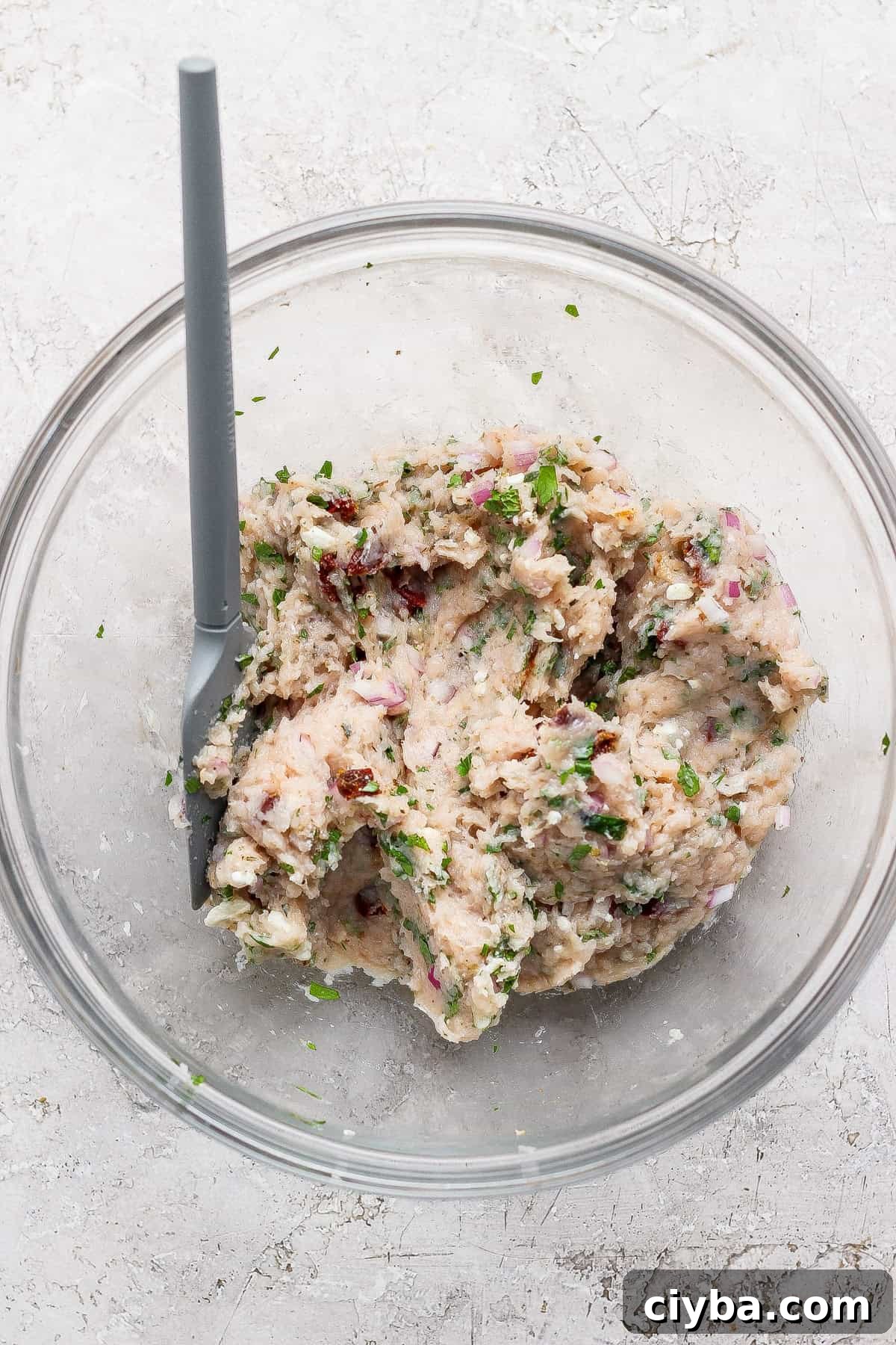 Crispy Air Fryer Mediterranean Chicken Bites with Herbs 4 A glass bowl with a gray spoon contains a mixture of mashed white beans, chopped herbs, red onion, and sun-dried tomatoes on a light-colored surface.