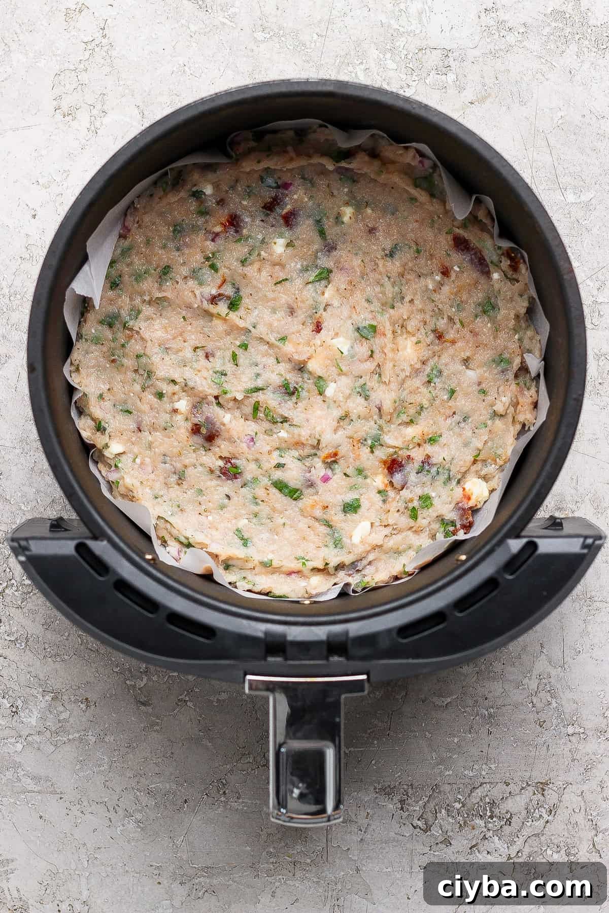 Crispy Air Fryer Mediterranean Chicken Bites with Herbs 5 Uncooked mixture of herbs, vegetables, and cheese pressed into a round baking dish lined with parchment paper inside an air fryer basket.
