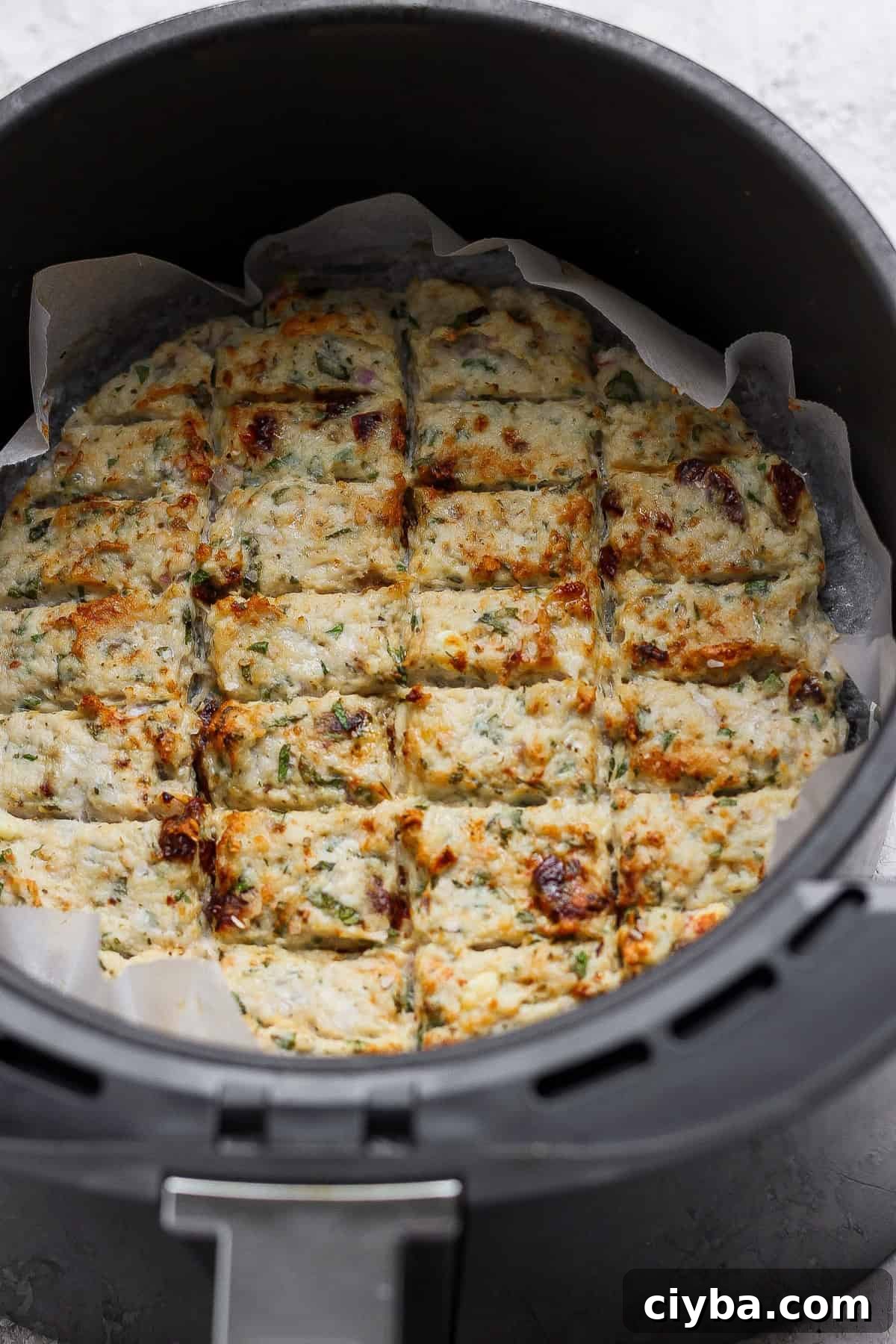 Crispy Air Fryer Mediterranean Chicken Bites with Herbs 6 A round dish of baked, sliced savory bars with herbs and browned spots, lined with parchment paper, inside an air fryer basket.