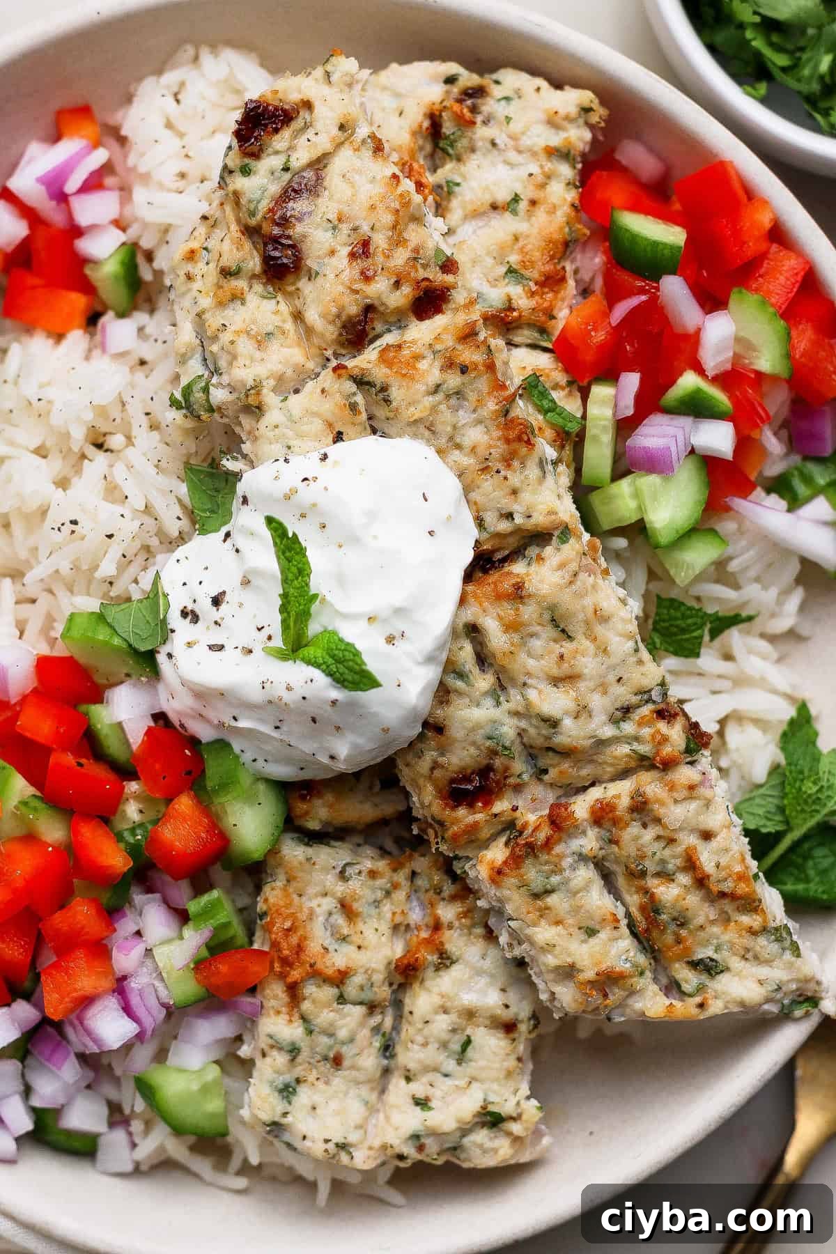 Crispy Air Fryer Mediterranean Chicken Bites with Herbs 7 A plate of rice topped with grilled chicken kebabs, chopped red onion, red bell pepper, cucumber, and a dollop of yogurt, garnished with a mint leaf.