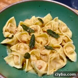 A green bowl filled with tortellini, topped with crispy sage and a sprinkle of cheese, placed on a wooden surface. The pasta is golden brown, glazed with a light sauce.
