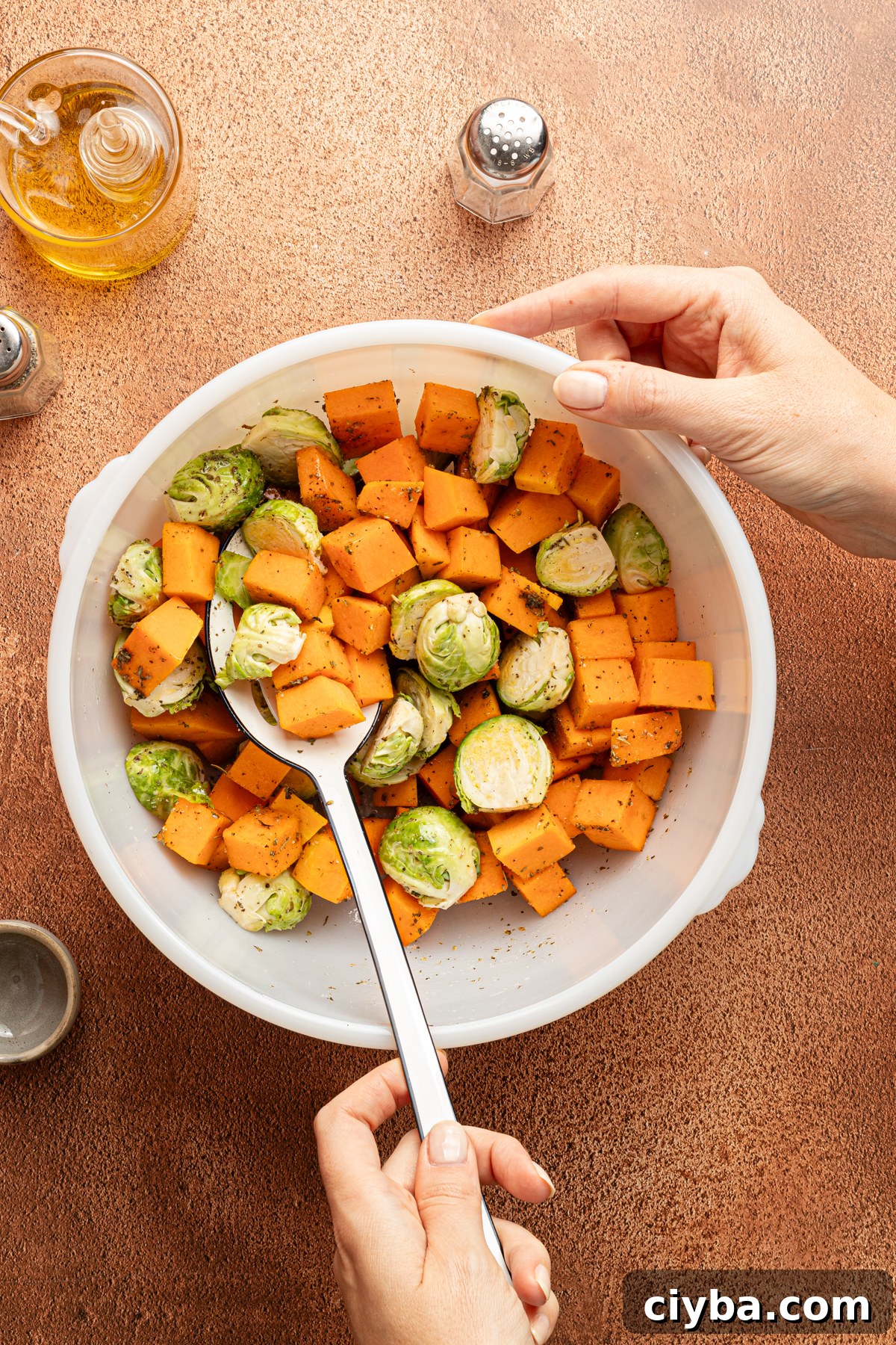 A large white mixing bowl filled with diced butternut squash and halved brussel sprouts being toseed with olive oil, Italian seasoning, salt and black pepper