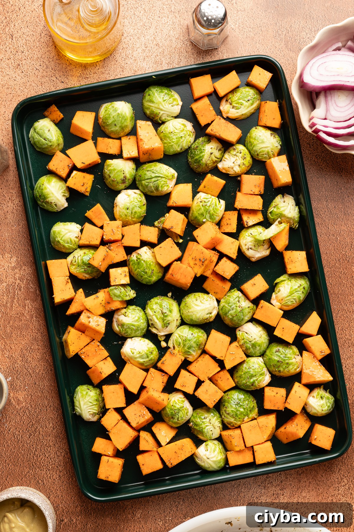 A dark green sheet pan filled with diced butternut squash and brussel sprouts tossed in aolive oil, Italian seasoning, salt and pepper