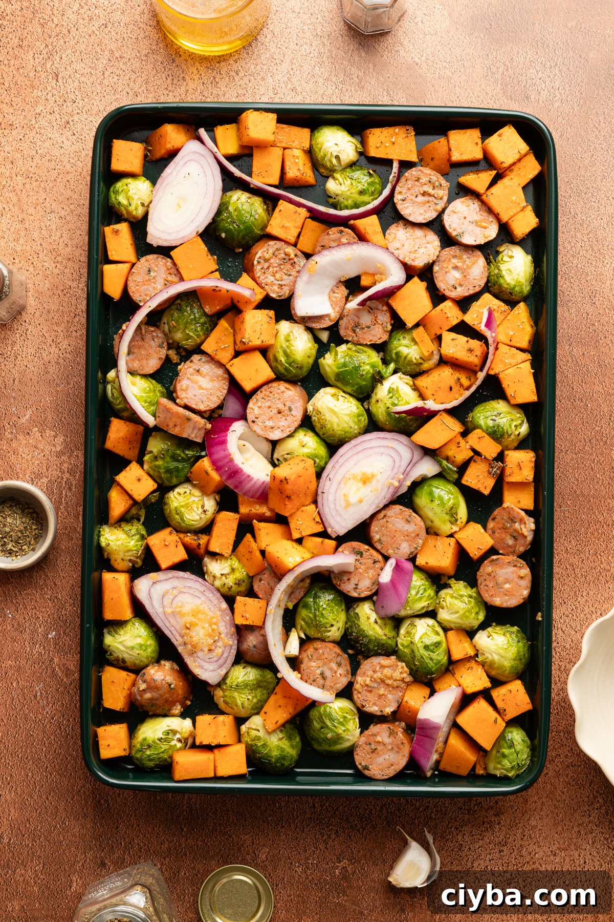 A dark green sheet pan filled with diced butternut squash, brussel sprouts, sliced red onion and chicken sausage