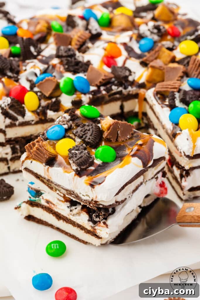 a square piece of ice cream sandwich cake being removed from the rest of the cake with a spatula. The slice has layers of ice cream sandwiches and cool whip, and is topped with candy and oreo pieces.