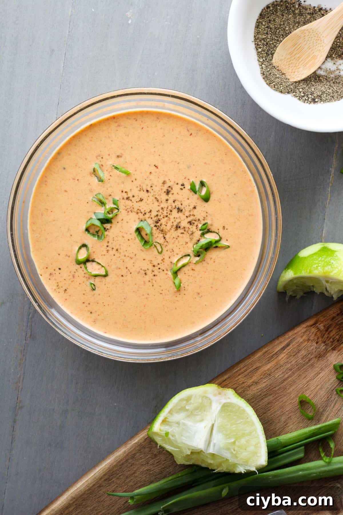 A small bowl of creamy chipotle lime sauce with a cutting board off center holding a lime and scallions.