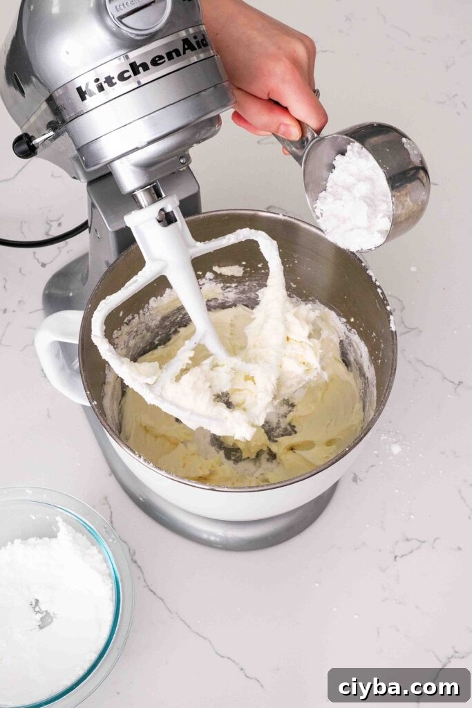 Red Velvet Cupcakes with Cream Cheese Frosting Recipe 10 A hand adds powdered sugar a cup at a time into a stand mixer bowl with butter.