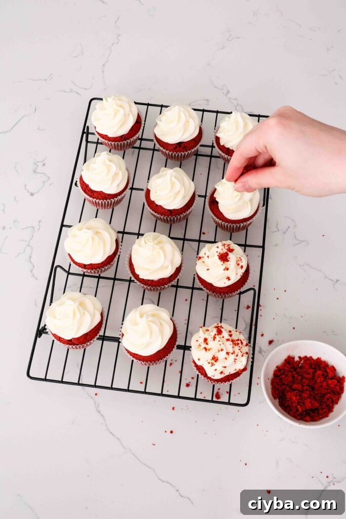 Red Velvet Cupcakes with Cream Cheese Frosting Recipe 15 A hand sprinkles red velvet crumbs on top of frosted red velvet cupcakes.