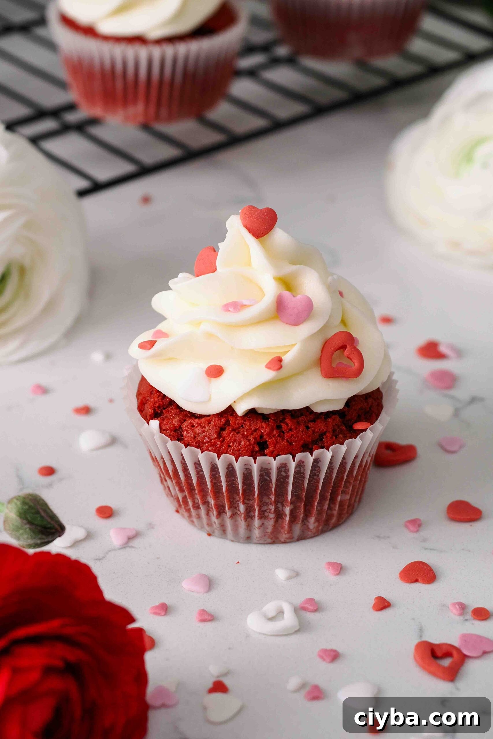 Red Velvet Cupcakes with Cream Cheese Frosting Recipe 16 A red velvet cupcake is topped with red, pink, and white heart sprinkles near a red ranunculus flower.