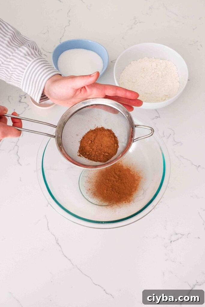 Red Velvet Cupcakes with Cream Cheese Frosting Recipe 3 Two hands sift cocoa powder into a glass bowl.