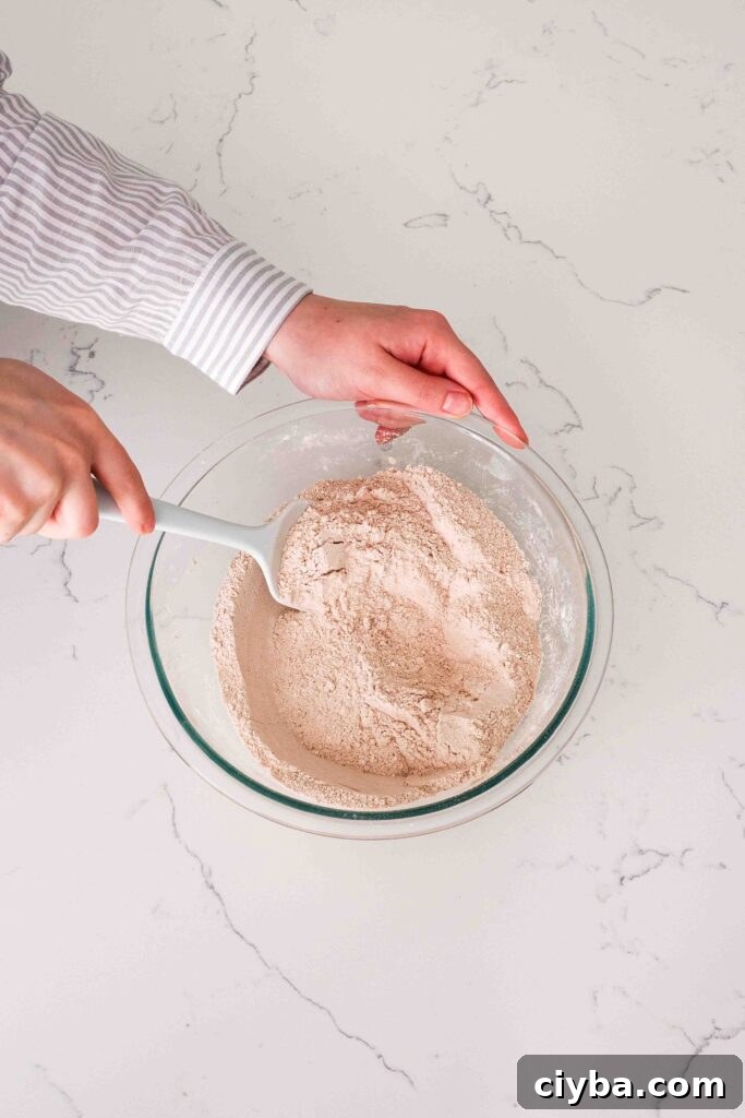 Red Velvet Cupcakes with Cream Cheese Frosting Recipe 4 Two hands use a spatula to stir cocoa powder into dry ingredients in a glass bowl.