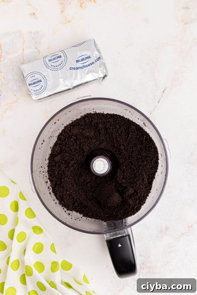 Crumbled Oreos in a food processor bowl with a package of cream cheese on a marble table. 