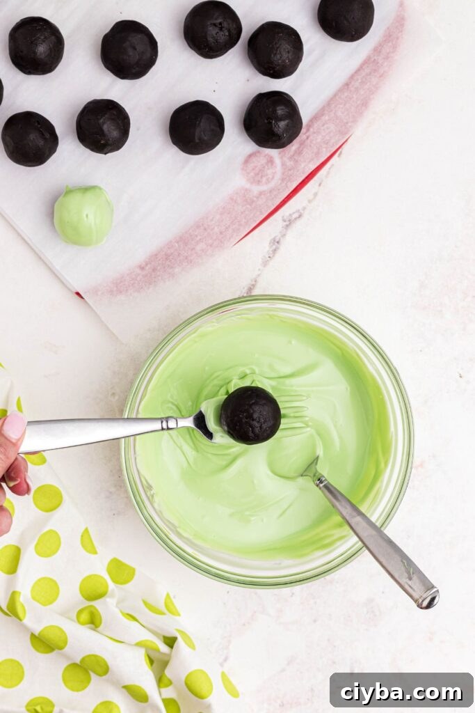 Oreo ball being dipped into melted green candy melts with remaining balls on parchment.