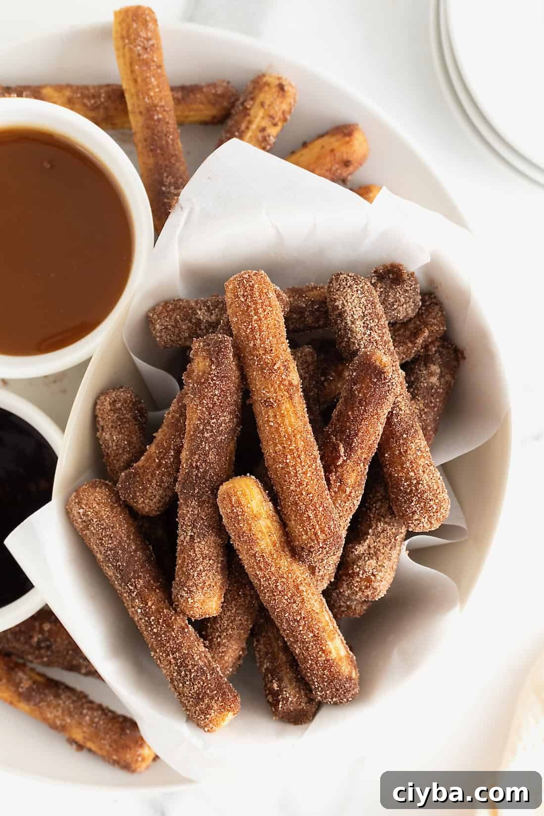 A parchment lined bowl of baked churros next to dipping sauces in white bowls.