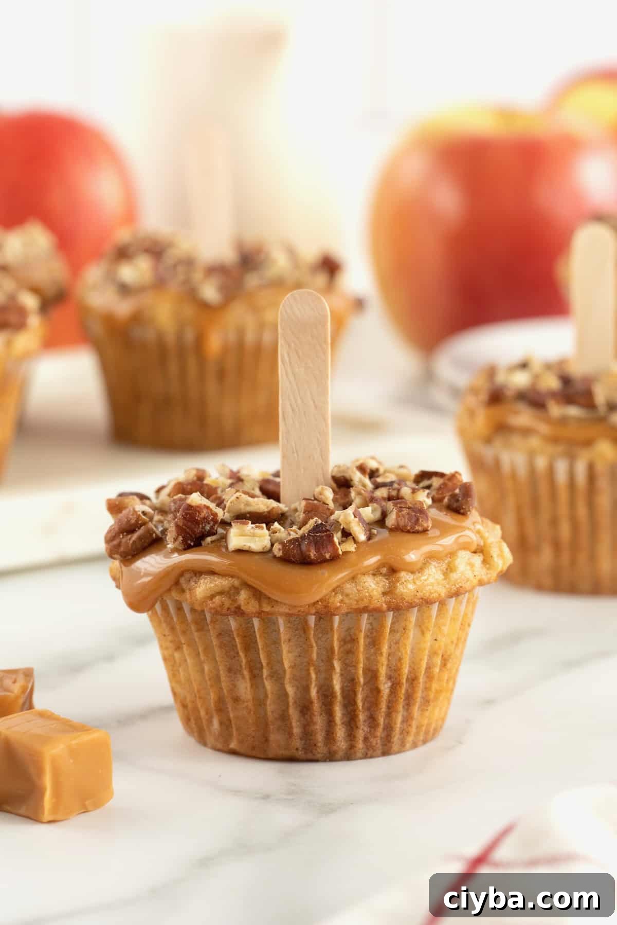 A caramel apple cupcake with caramel and pecan pieces on top and a popsicle stick in the center.