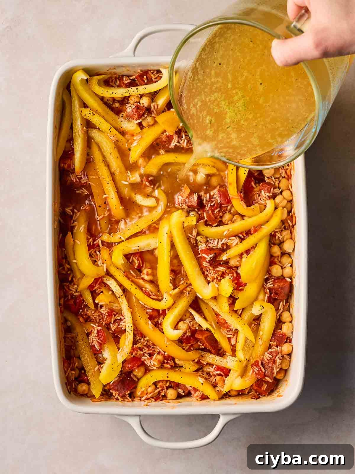 One-Pan Chorizo and Rice Bake: Dump, Bake, Serve 7 A hand pouring stock from a jug into an oven dish with lot of ingredients, including chorizo, rice, peppers and chickpeas.