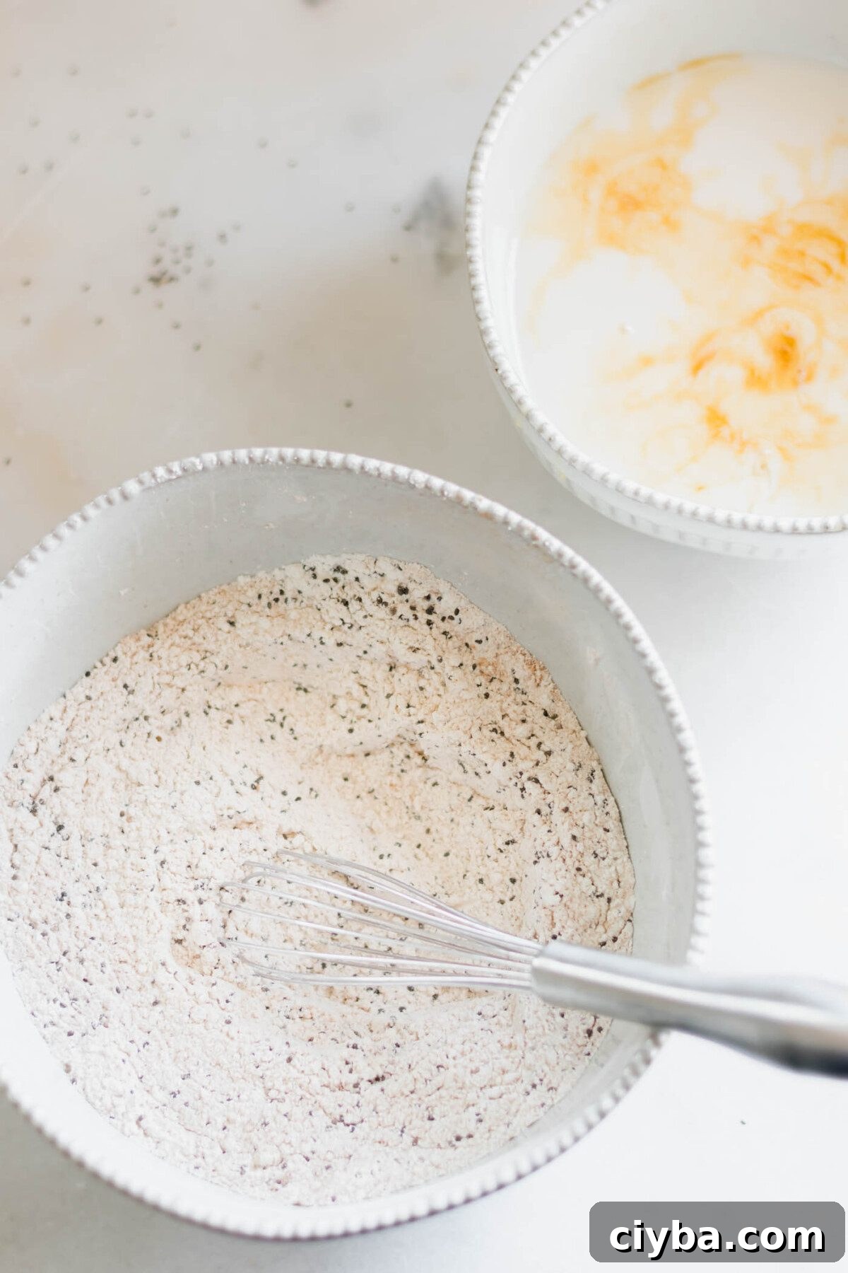 One bowl with dry pancake ingredients and a whisk in it and one bowl with milk, vanilla and vinegar.